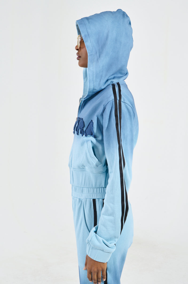 Against a plain background, a person is in profile wearing the "DON'T TALK TO ME I'M RUNNING ERRANDS ZIP UP SWEATSHIRT" in light blue with black striped sleeves. Styled with short hair and sunglasses, their hands are comfortably tucked into the pockets, exuding a sporty yet relaxed vibe.