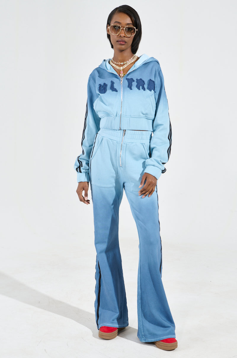 Dressed in the DON'T TALK TO ME I'M RUNNING ERRANDS ZIP UP SWEATSHIRT, a person stands against a plain background. They pair the chic ombré blue zip-up hoodie with matching flared pants and accessorize with stylish sunglasses, layered necklaces, and red shoes.