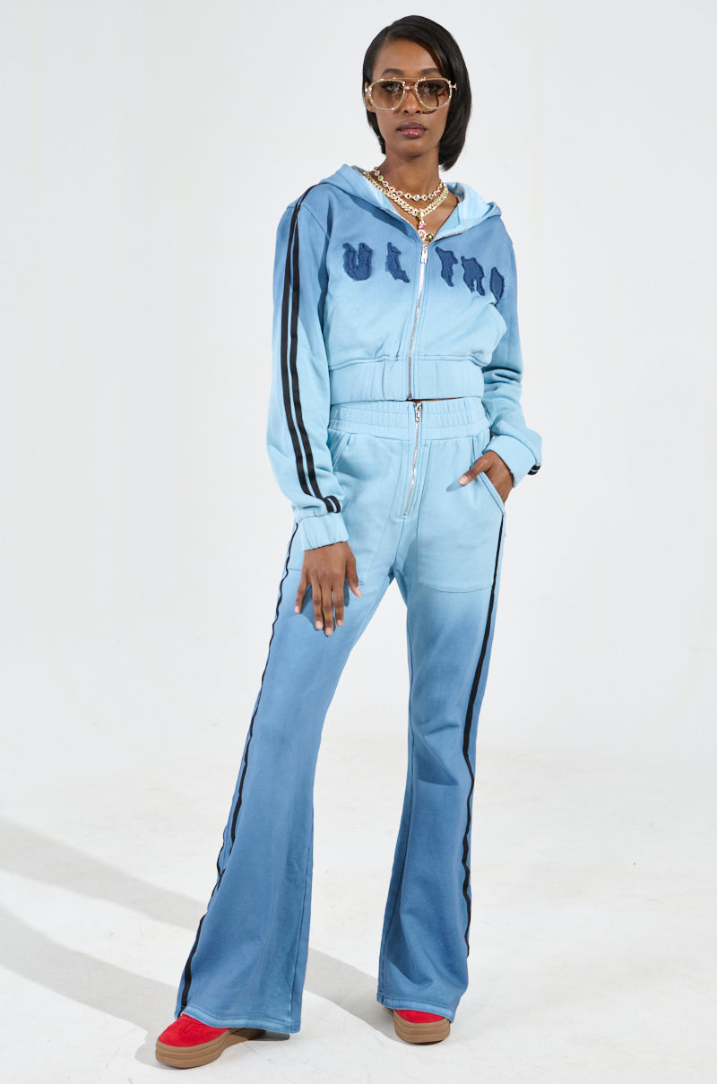 A person poses confidently in the DON'T TALK TO ME I'M RUNNING ERRANDS ZIP UP SWEATSHIRT, featuring an ombré light blue color with black side stripes. Paired with red shoes and complemented by sunglasses, a gold necklace, and the hood up, they create a bold look. The cropped silhouette adds flair against the plain white background.