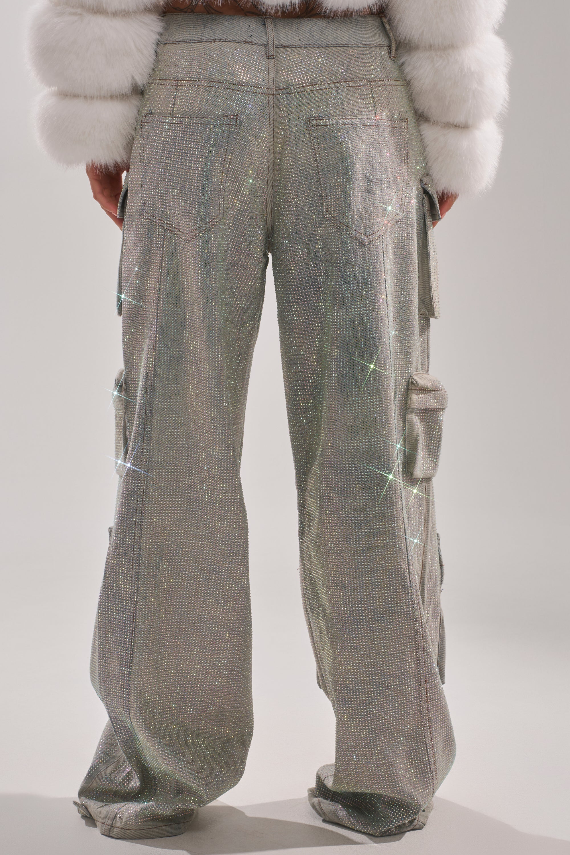A person is seen from behind wearing the BLING BLING CARGO DENIM—shimmery, silver high-rise cargo pants with sparkling accents and pockets—paired with a white furry jacket, showcasing the pants' unique texture and eye-catching details. 3