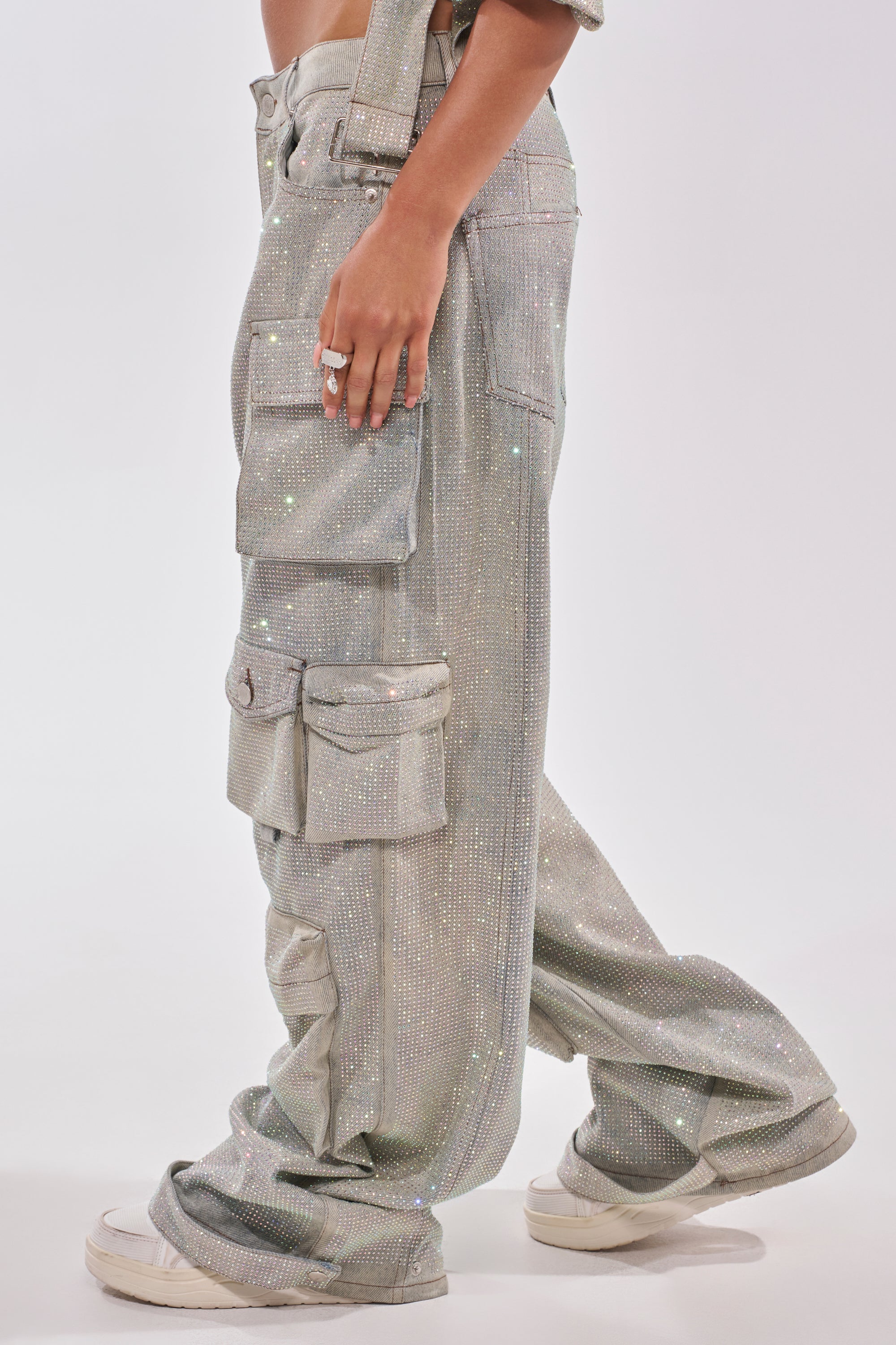 Model wears the BLING BLING CARGO DENIM—loose, sparkling silver high-rise cargo pants with large pockets—paired with white sneakers, shown from midriff to feet against a plain white background. 3