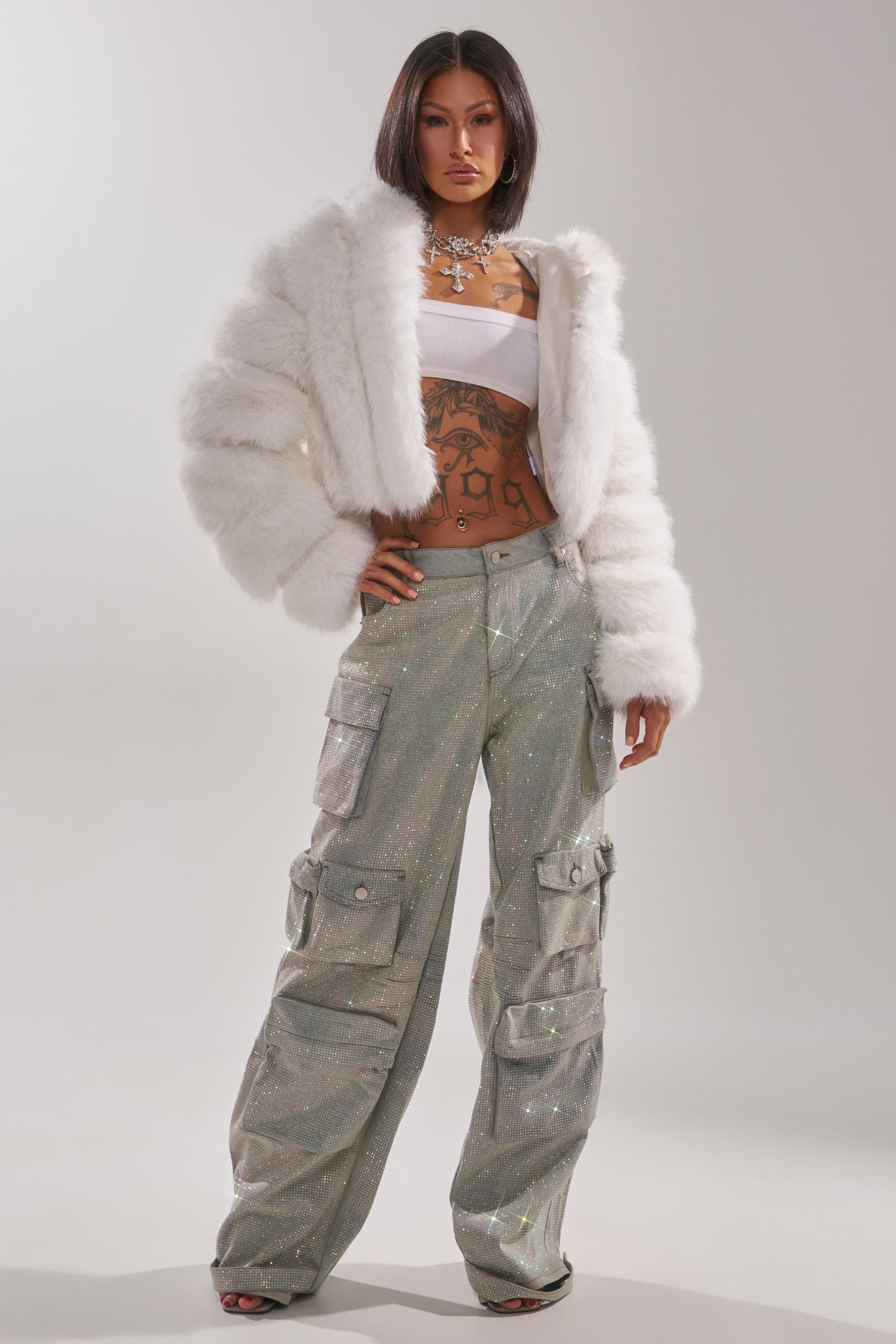 A woman poses confidently in BLING BLING CARGO DENIM with a wide-leg fit, paired with a white faux fur jacket and bandeau top. She accessorizes with bold jewelry and displays midriff tattoos against a plain, light background. 4