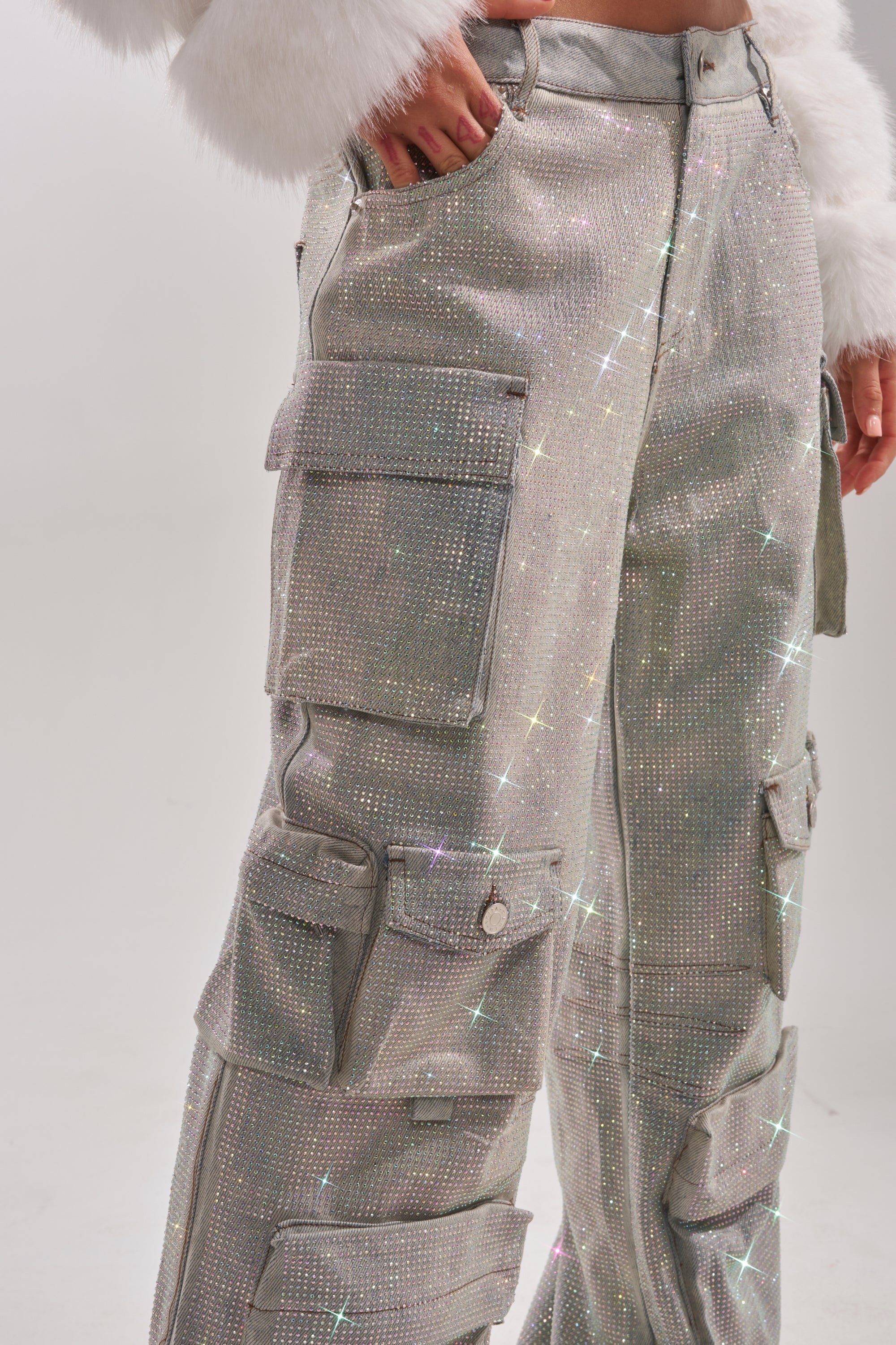 Someone stands against a plain light background wearing the BLING BLING CARGO DENIM—sparkly silver cargo pants with a wide-leg silhouette and multiple pockets—paired with a white fuzzy jacket. 7