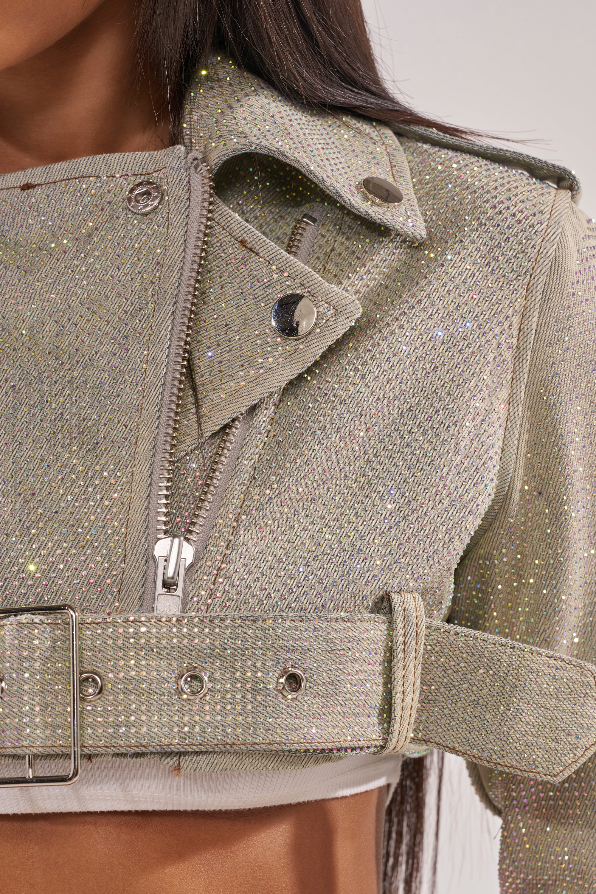 Close-up of a person wearing the SHINE BRIGHT DENIM MOTO jacket featuring rhinestones, sparkling texture, metallic buttons, zipper, and belt; only lower face, hair, neck, and upper torso are visible. 10