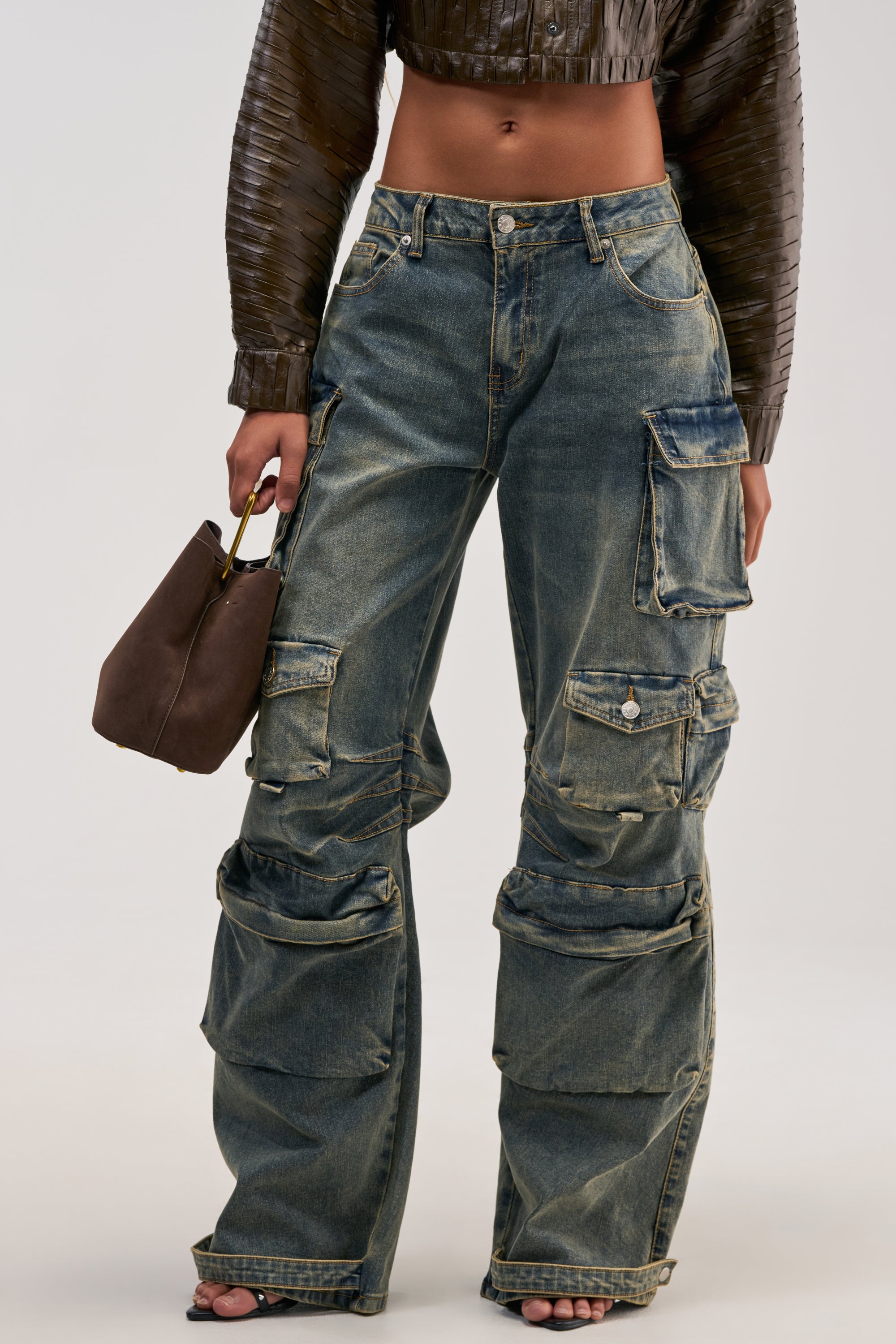A person wearing the VINTAGE LOVER CARGO DENIM, featuring a wide-leg silhouette and multiple pockets, styled with a cropped brown textured jacket and holding a brown handbag. The image is cropped from shoulders to feet, effortlessly highlighting a modern streetwear vibe.