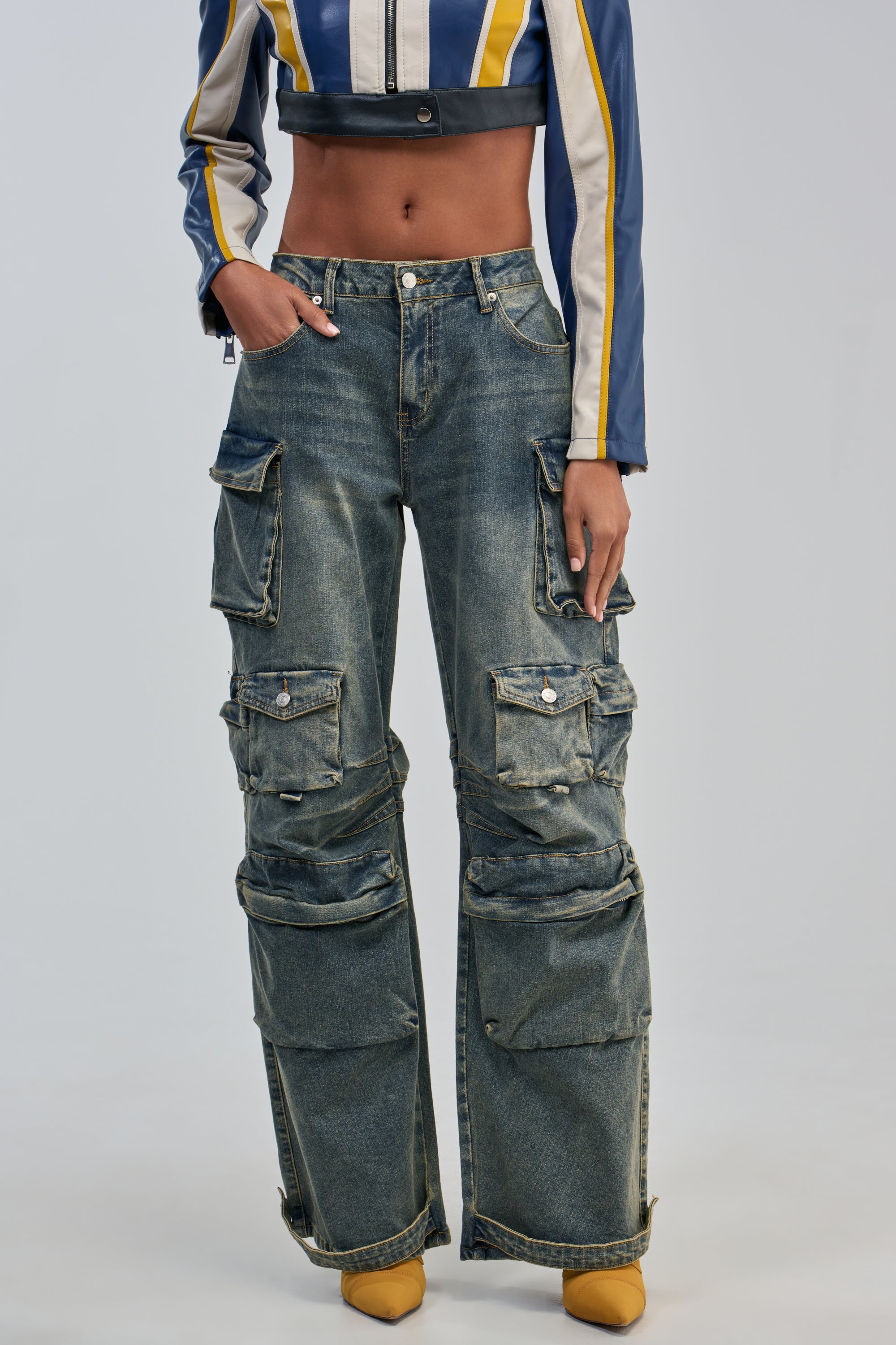 Model stands against a plain background, showcasing an effortlessly cool streetwear look featuring the VINTAGE LOVER CARGO DENIM—a pair of vintage-washed, wide-leg cargo jeans with multiple pockets—styled with a cropped jacket and yellow pointed-toe shoes.
