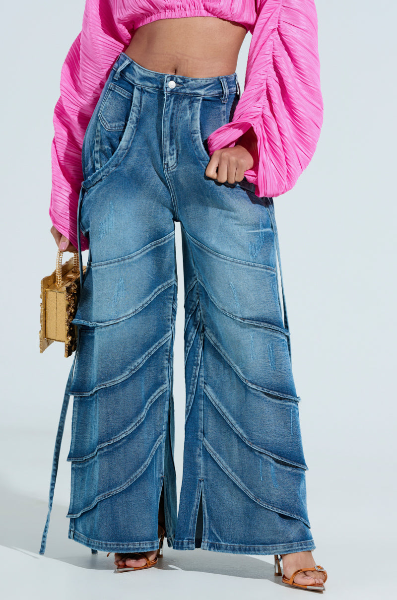 Wearing RIDE THE WAVE WIDE LEG DENIM JEANS with flared design and a pink textured long-sleeve top, they hold a small gold purse and sport open-toed heels, achieving a bold and stylish look.