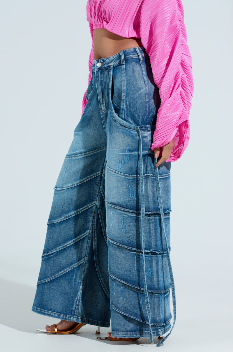 A person wears the RIDE THE WAVE WIDE LEG DENIM JEANS, featuring distressed blue denim with unique seam detailing, paired with a bright pink pleated top and open-toed shoes. The high-rise jeans enhance the ensemble, highlighting the textures and style of the outfit.