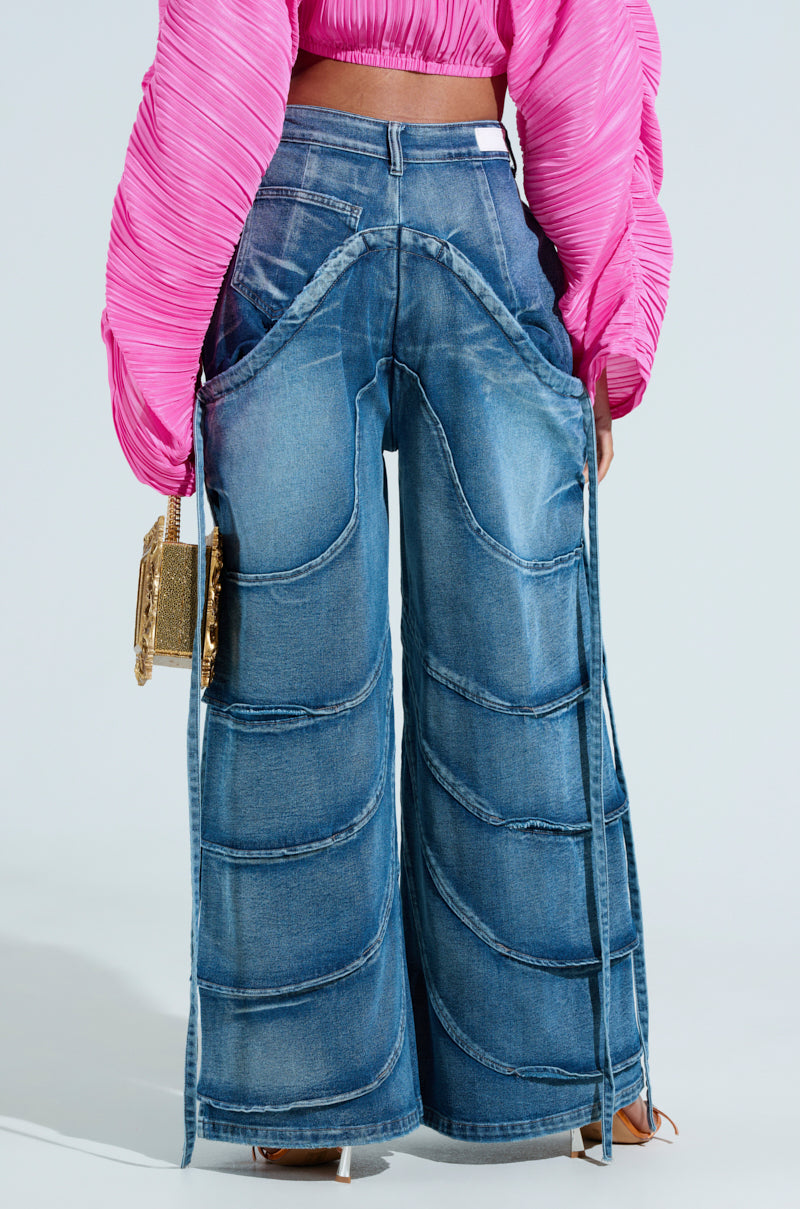 Someone in RIDE THE WAVE WIDE LEG DENIM JEANS with curved stitching and a bright pink textured top holds a small gold purse. A tag peeks from the jeans' waistband, contrasting against the light background.