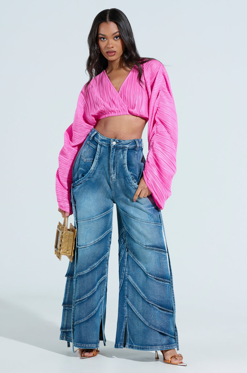 A person with long hair sports a vibrant pink, long-sleeved crop top and RIDE THE WAVE WIDE LEG DENIM JEANS, holding a small square handbag while standing confidently against a plain background.