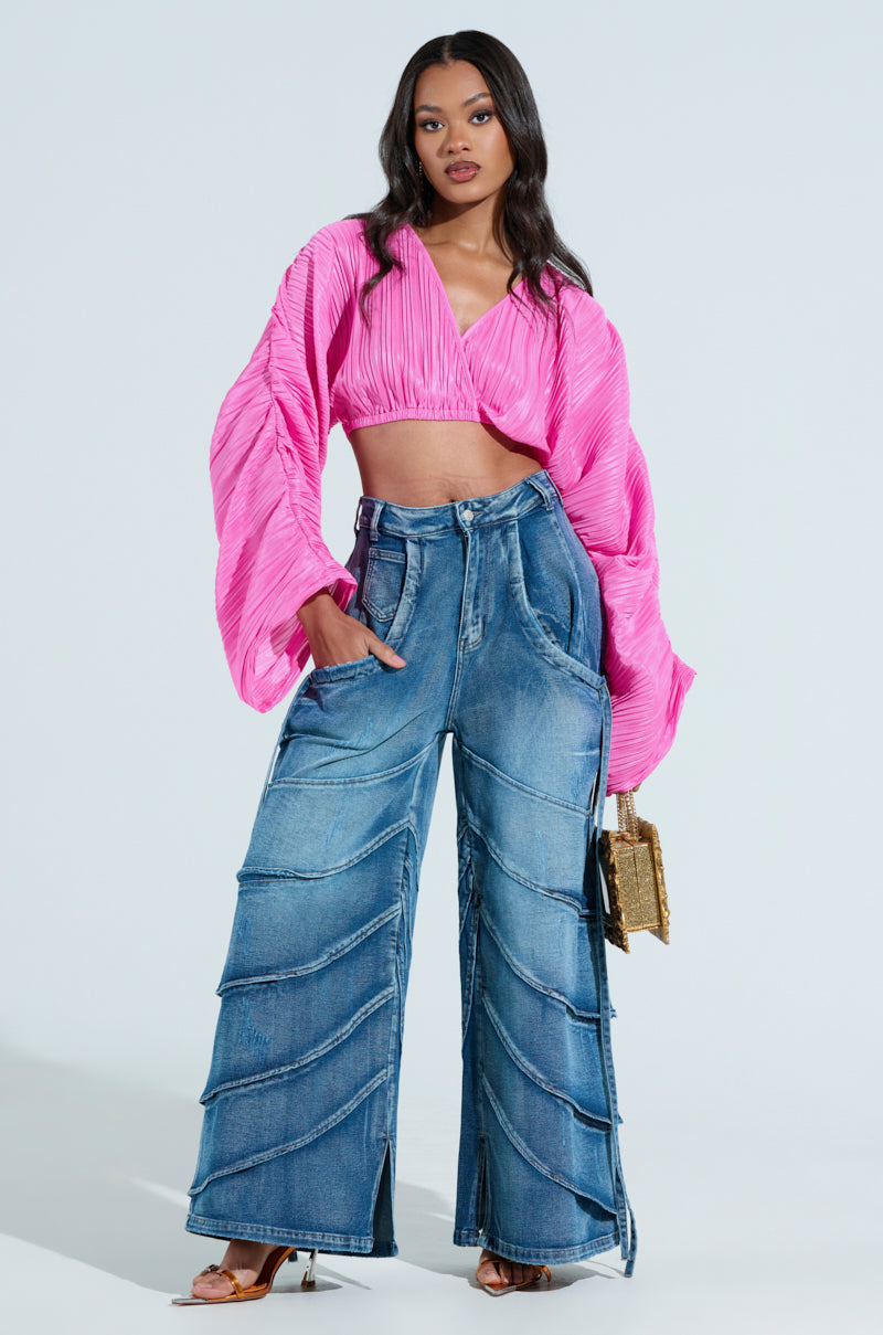A woman poses in a bright pink pleated crop top with wide sleeves, paired with RIDE THE WAVE WIDE LEG DENIM JEANS. She accessorizes with a gold clutch and tan heels against a simple white backdrop.