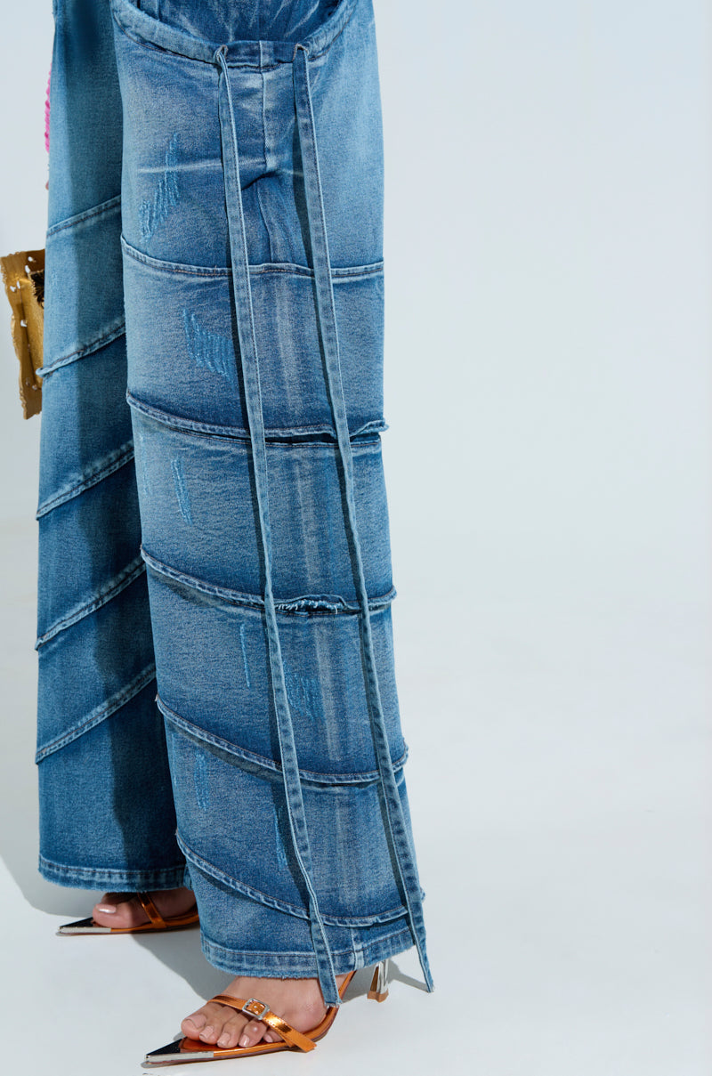 Close-up of a person in RIDE THE WAVE WIDE LEG DENIM JEANS, which feature layered horizontal seams and a high-rise flared design with long cascading straps. The look is finished with pointed, strappy heels in shimmering metallic tones.