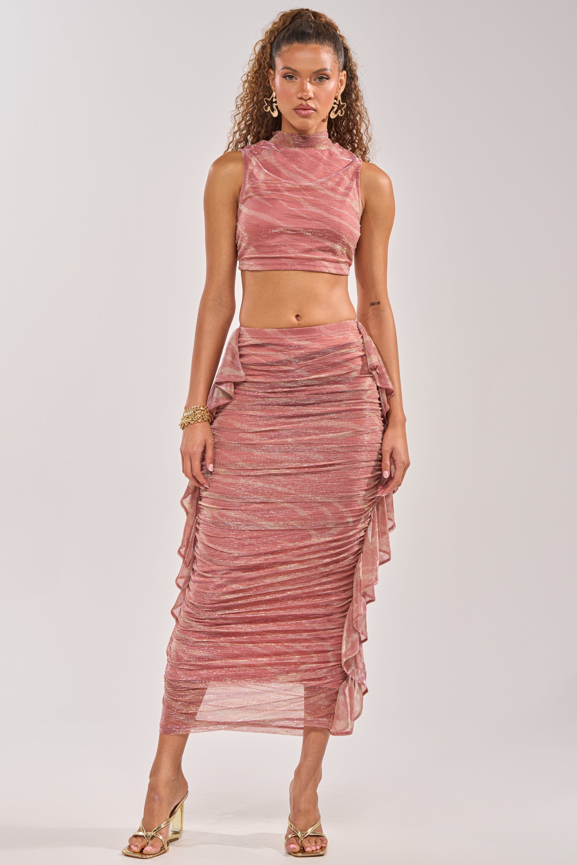A woman wears the PLAY WITH ME MAXI SKIRT—a high-waisted pink metallic skirt with ruffle and ruched sides. She pairs it with a sleeveless crop top, curly hair, gold hoop earrings, bracelets, and gold heeled sandals for a chic vacation look. 0