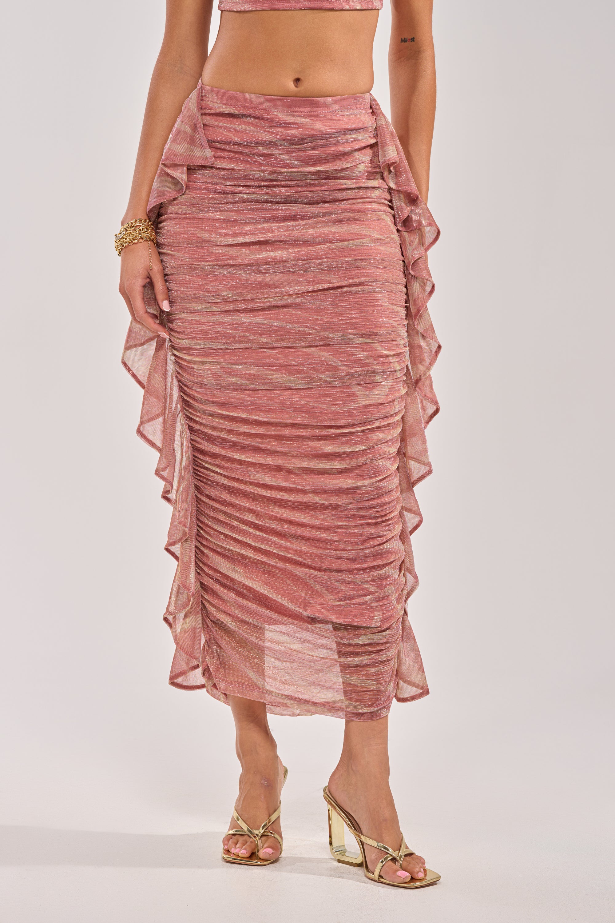 A woman models the PLAY WITH ME MAXI SKIRT, a pink marbled maxi skirt with ruffle side details, paired with a matching crop top and gold accessories—perfect vacation wear shown against a white background. 1