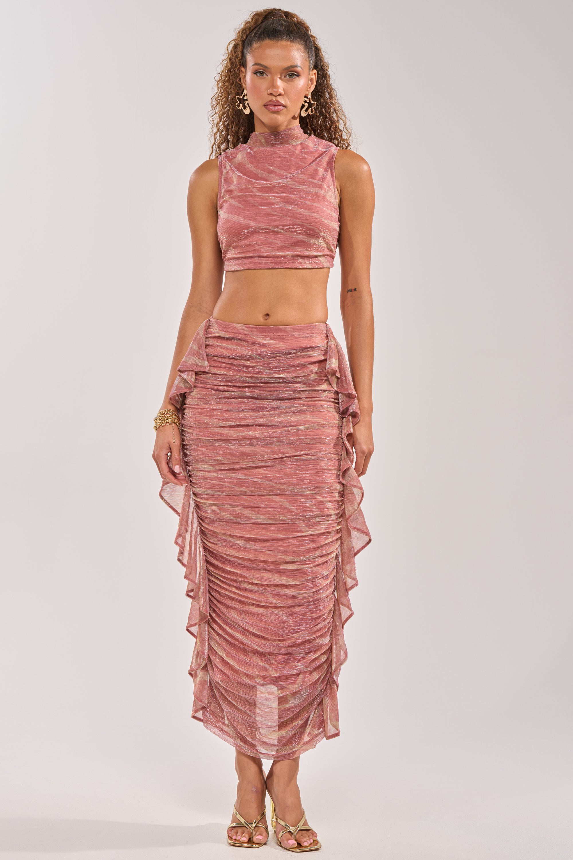 A woman models the PLAY WITH ME MAXI SKIRT—a pink, ruched, ruffle-detail maxi skirt—paired with a matching sleeveless high-neck crop top, gold hoop earrings, bracelets, and gold heeled sandals against a light gray background. 5