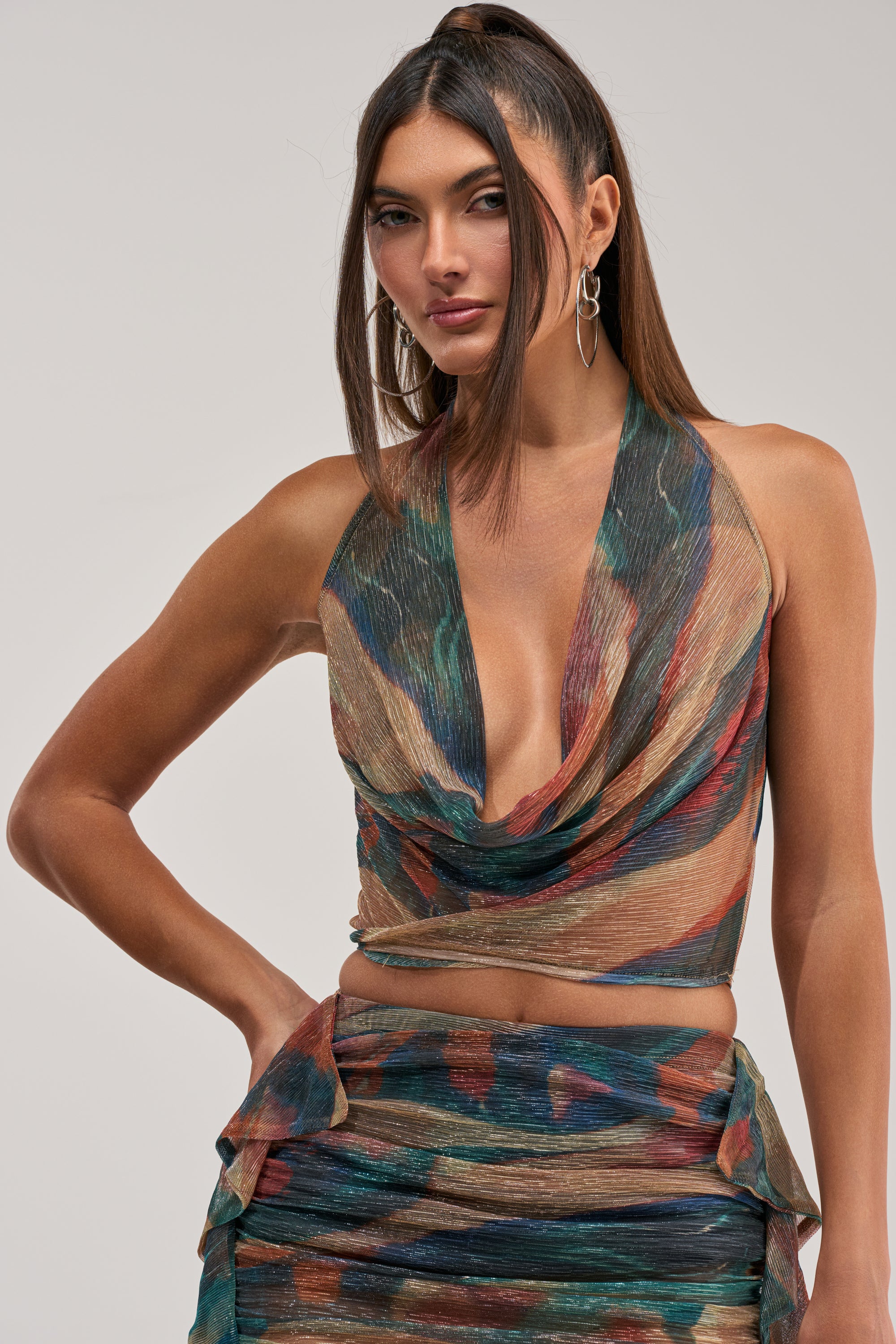 A woman wears the DROP IT LOW COWL NECK BLOUSE—a sleeveless, multicolored halter crop top—paired with a matching skirt, posing confidently with one hand on her hip against a plain background; perfect for any night out.
