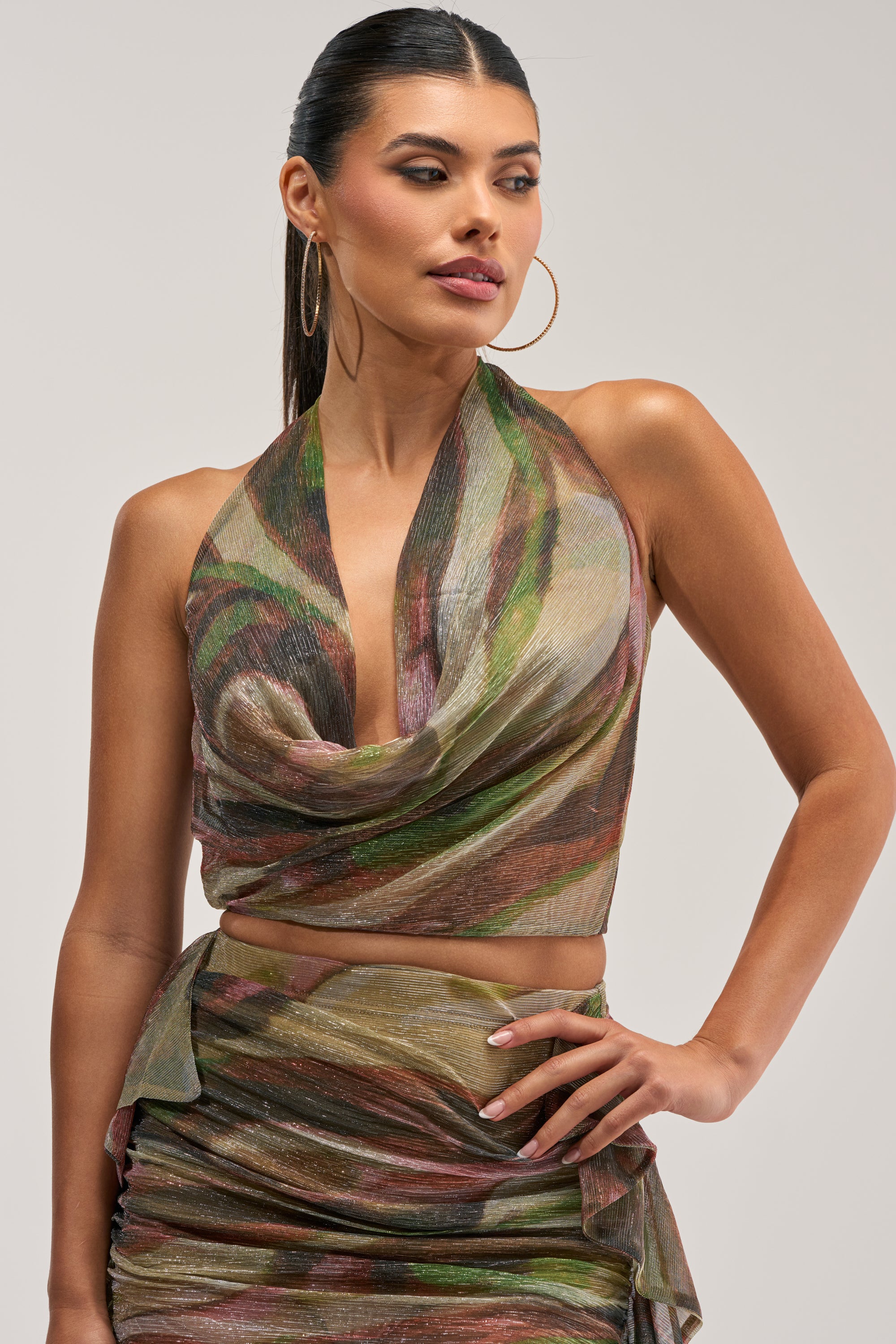 A woman with long dark hair styled in a sleek ponytail wears the DROP IT LOW COWL NECK BLOUSE IN GREEN MULTI, a sleeveless halter top featuring an abstract print and cowl neckline, paired with a matching skirt. Hoop earrings complete her effortlessly chic, vacation-ready look as she gazes to her left against a plain background.