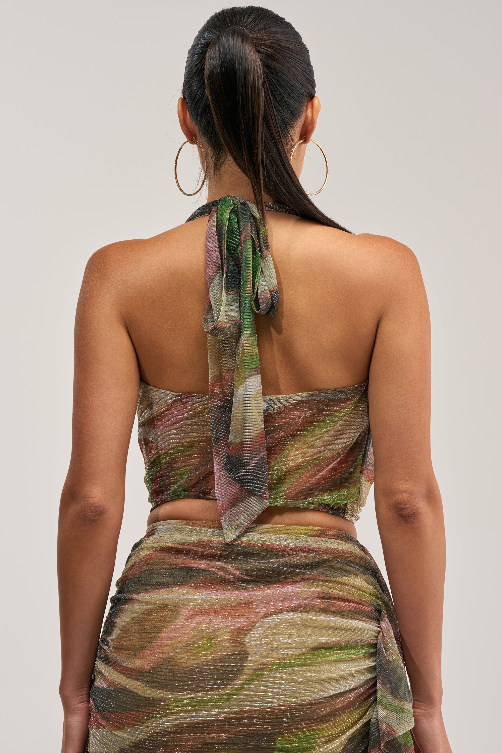 A woman with long, dark hair in a ponytail is seen from behind wearing the DROP IT LOW COWL NECK BLOUSE IN GREEN MULTI, featuring an abstract swirl pattern in green, pink, and brown. The back tie detail of her cowl neck halter blouse and her large hoop earrings complete her effortlessly vacation-ready look.