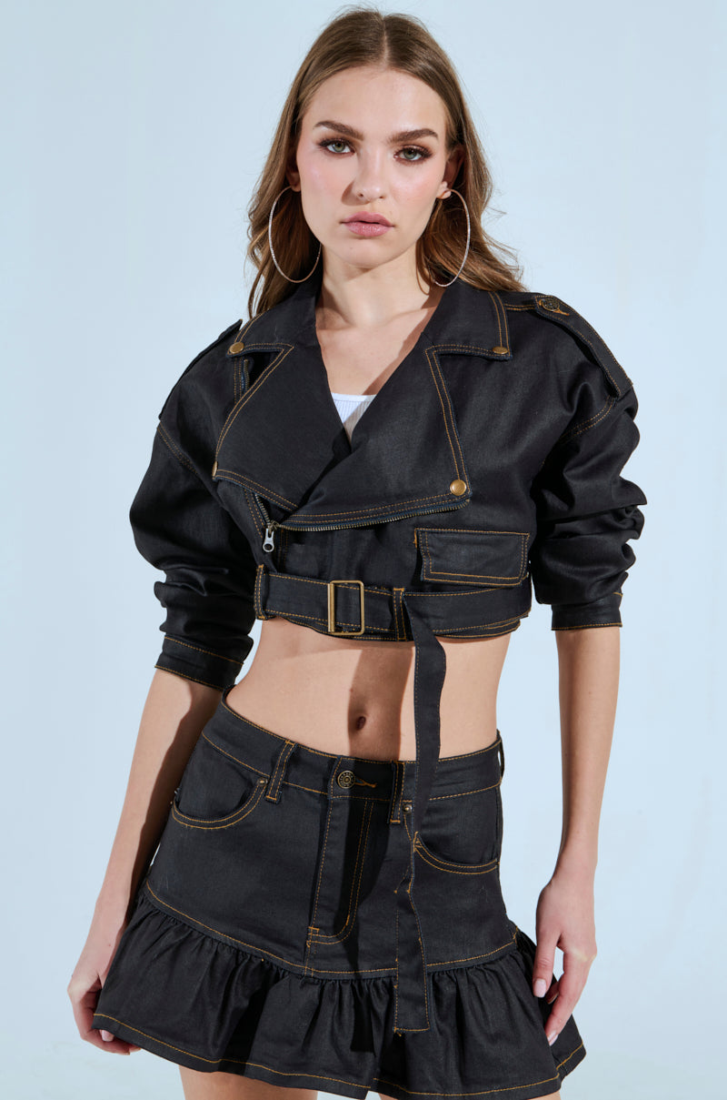 Against a light blue background, a woman wears the BETTER DAYS AHEAD COATED DENIM MOTO jacket and matching high-waisted skirt. She has long hair, hoop earrings, and exudes confidence.