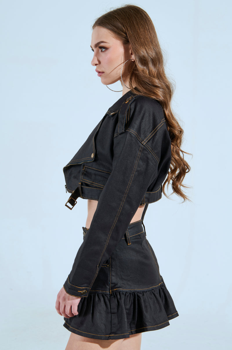 A woman with long hair is wearing the BETTER DAYS AHEAD COATED DENIM MOTO in black and a matching ruffled skirt, gazing sideways against a plain light blue background.