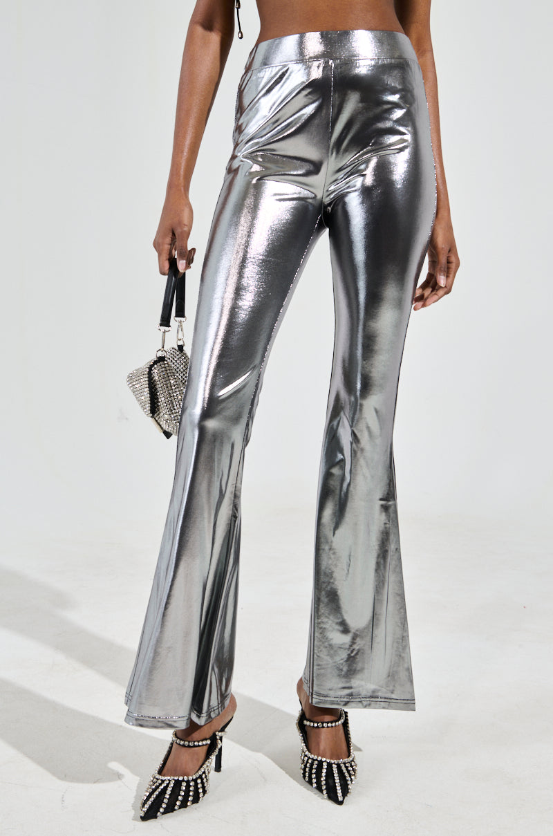 A person wearing MIAMI NIGHTS RUCHED LEGGING in shiny metallic silver and black studded heels holds a small silver and black handbag, with a plain gray background highlighting the party-perfect look of the outfit.