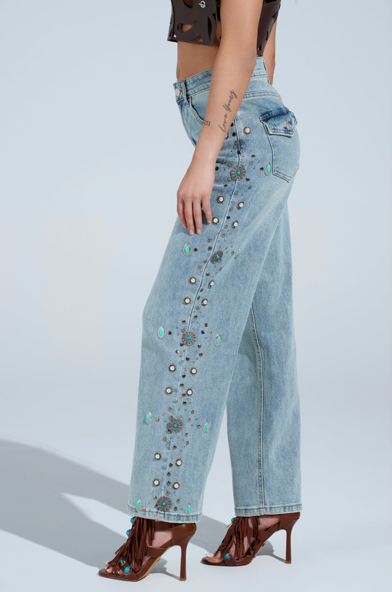 A person in WESTERN BABY DENIM PANTS—light blue, high-waisted cowboy jeans with floral and pearl embellishments—paired with brown heeled sandals featuring fringe and a sleeveless top, stands against a plain background.