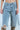 A person dons ALL IVE GOT DENIM BERMUDA SHORTS, featuring distressed detailing, large rips, and a detachable chain on the pocket, teamed with knee-high snakeskin-patterned boots. The image focuses on the lower half against a light blue background.