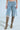 Someone flaunts the ALL IVE GOT DENIM BERMUDA SHORT, featuring distressed details and a detachable chain, paired with knee-high snakeskin boots. The trendy shorts display thigh tears, while the minimalist backdrop highlights this fashion-forward ensemble.