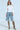 A person stands confidently wearing sunglasses, a white open-shoulder blouse, and ALL IVE GOT DENIM BERMUDA SHORT. The outfit is paired with high snake-patterned boots and a detachable chain attachment, set against a plain background.
