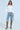 A person confidently wears a white cold-shoulder button-up shirt, ALL IVE GOT DENIM BERMUDA SHORT with distressed detailing, sunglasses, and high-heeled patterned boots against a plain light-colored background.