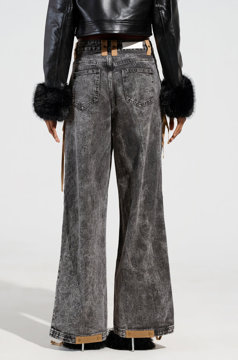 A person wearing the LACED WITH STYLE LACE UP DENIM JEANS in black, featuring high-rise wide legs and distressed details, paired with a leather belt and a cropped black leather jacket with fur cuffs, viewed from behind.