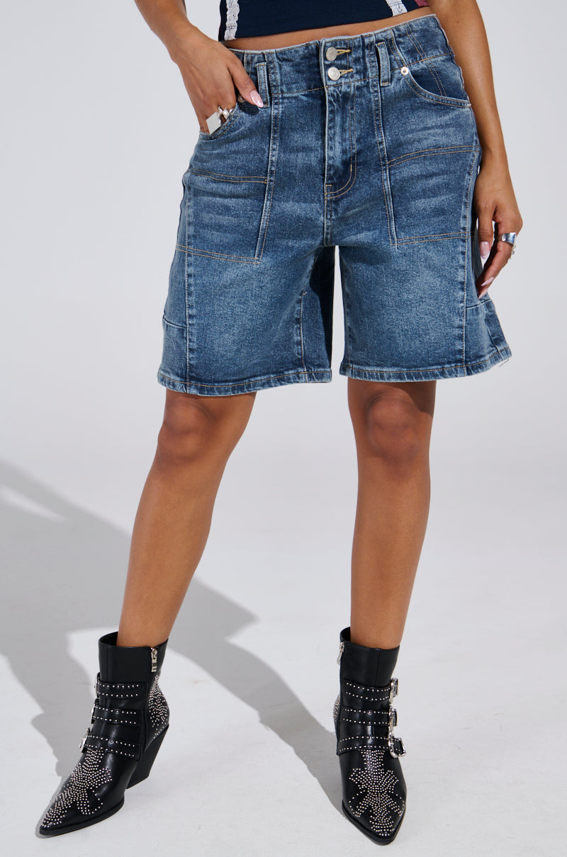 Wearing a blue sleeveless top and TIME IS TICKING BERMUDA SHORT, paired with black studded ankle boots, the person exudes a relaxed vibe in the loose-leg fit as they stand with hands in pockets, showcasing their casual style.