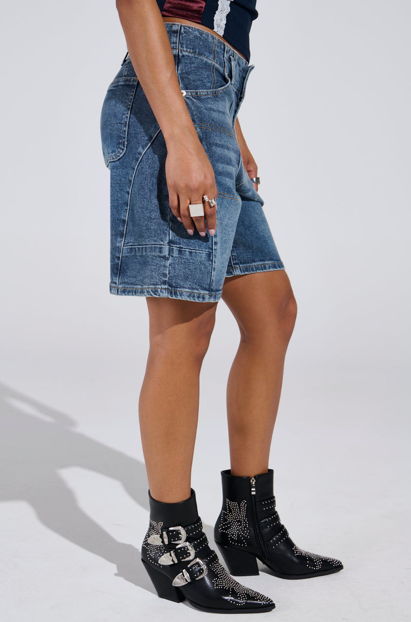The individual wears TIME IS TICKING BERMUDA SHORTS, studded black ankle boots with buckles, and a dark sleeveless top. Their hands feature several rings against a plain light grey background.
