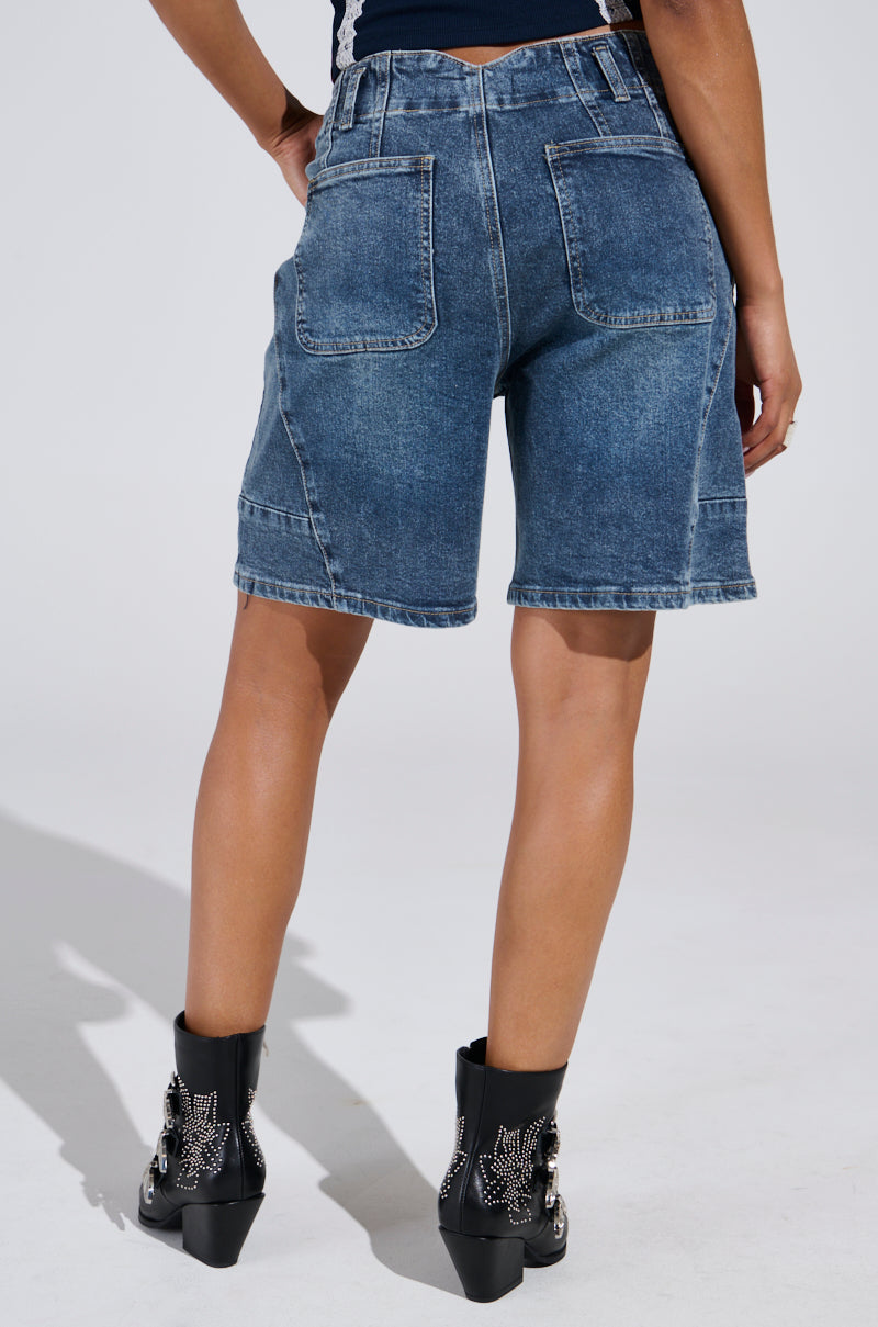 The person is shown from the back wearing TIME IS TICKING BERMUDA SHORTS and loose-leg knee-length fit, paired with black studded ankle boots against a plain, light-colored background.