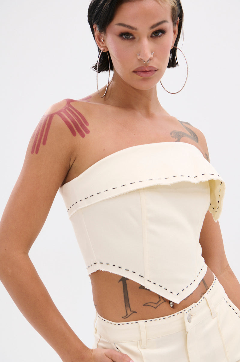 A person with short dark hair and hoop earrings wears the BREAK MY LOVE LINEN TUBE TOP, a cream-colored piece featuring an asymmetric neckline and visible stitching. Their tattoos include a hand on their shoulder and various torso designs. 2