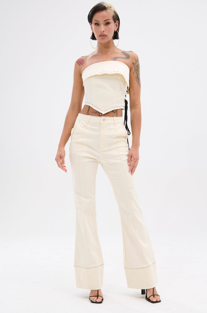 A woman with short dark hair, visible tattoos, and black heeled sandals faces forward wearing the BREAK MY LOVE LINEN TUBE TOP—an off-white linen, asymmetric tube top with studded trim—paired with matching high-waisted flared pants. 7