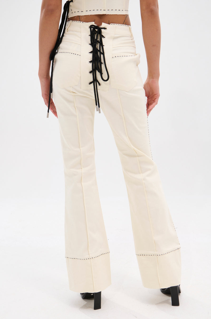 A person stands with their back turned, wearing the BREAK MY LOVE LINEN LACE UP PANT in cream with a flared silhouette, visible stitching, and black heels. The high-rise fit flatters against a plain white background.
