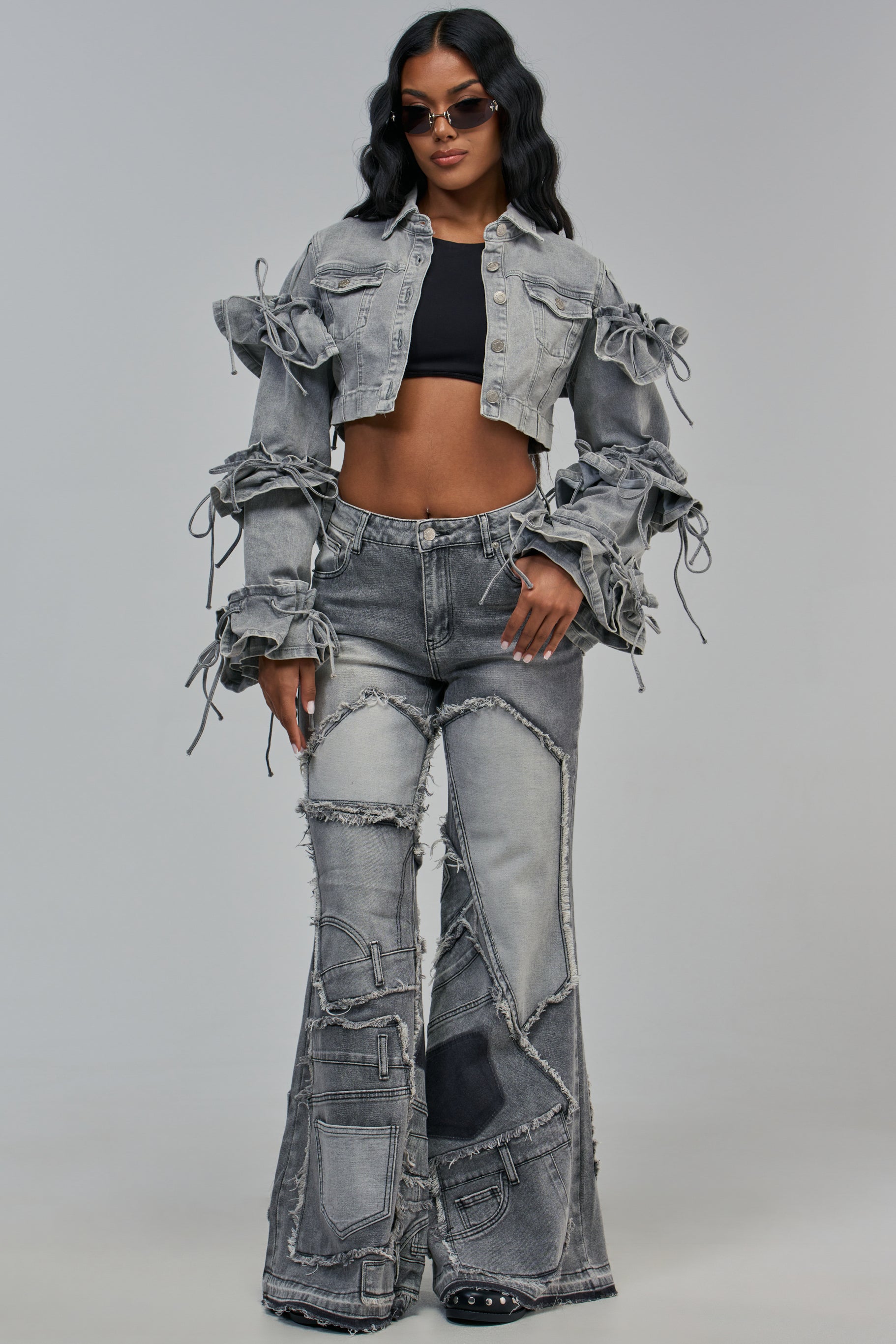 A person wearing a grey denim cropped jacket with ruffled sleeves and matching ALL OVER ME DENIM PATCHWORK JEANS IN GREY MULTI in a trendy streetwear style.