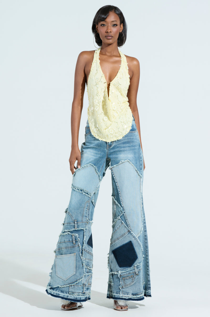 A woman wears ALL OVER ME DENIM PATCHWORK JEANS with a yellow lace halter top, confidently displaying the flared leg silhouette against a light background in shades of denim.
