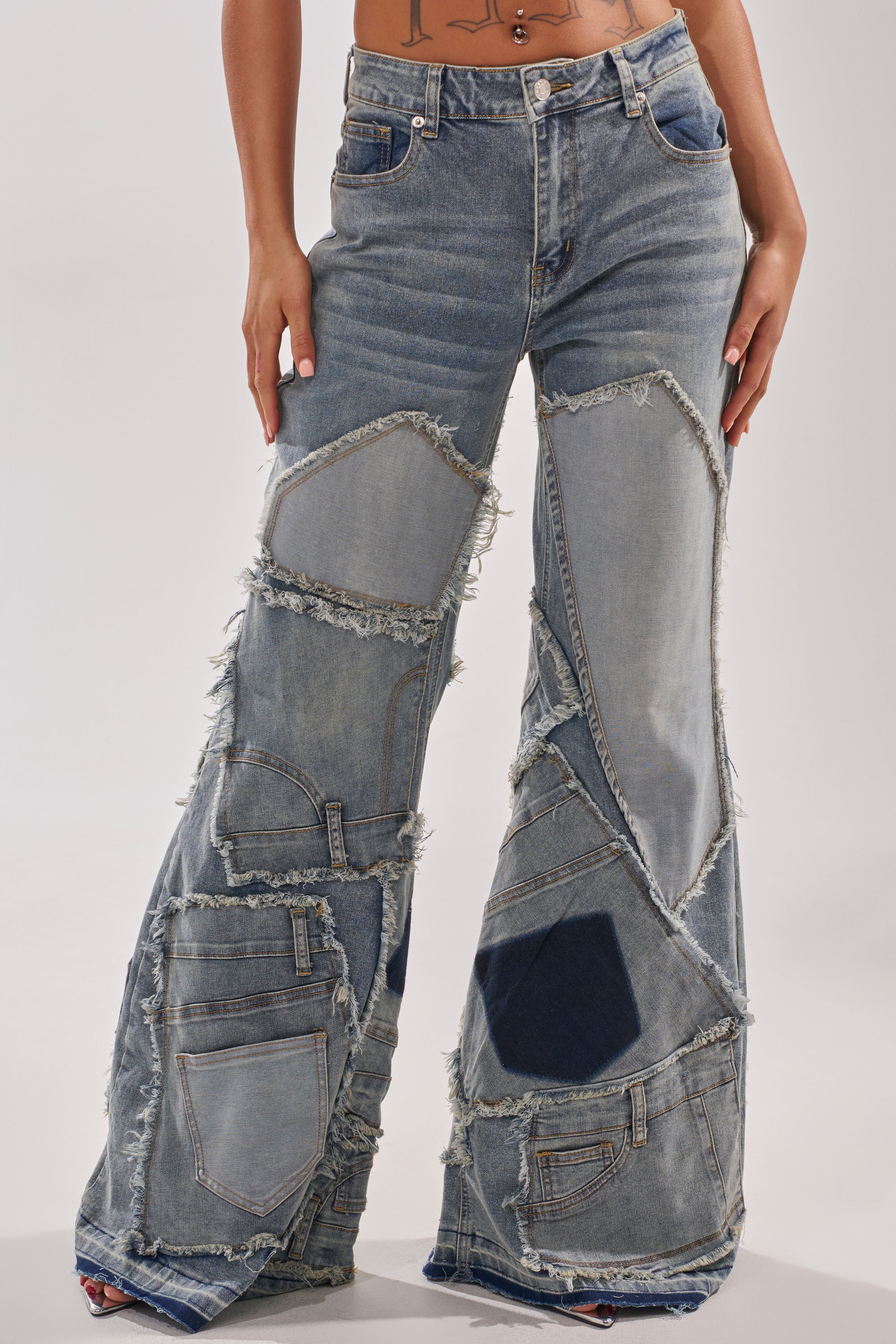 Wearing the ALL OVER ME DENIM PATCHWORK JEANS, a person shows off high-rise, wide-leg blue jeans featuring frayed patchwork details, mixed denim textures, multiple pockets, and a flared leg for an edgy look. Their hands rest at their sides.