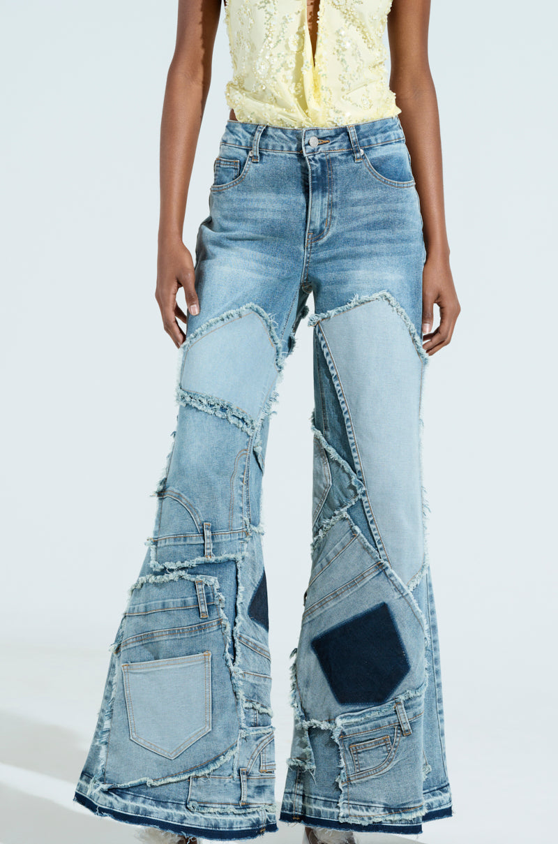 Wearing the "ALL OVER ME DENIM PATCHWORK JEANS," a person confidently showcases high-rise, flared blue denim bell-bottoms with frayed edges and textured patches. Paired with a white floral top, these unique jeans stand out against the plain background.