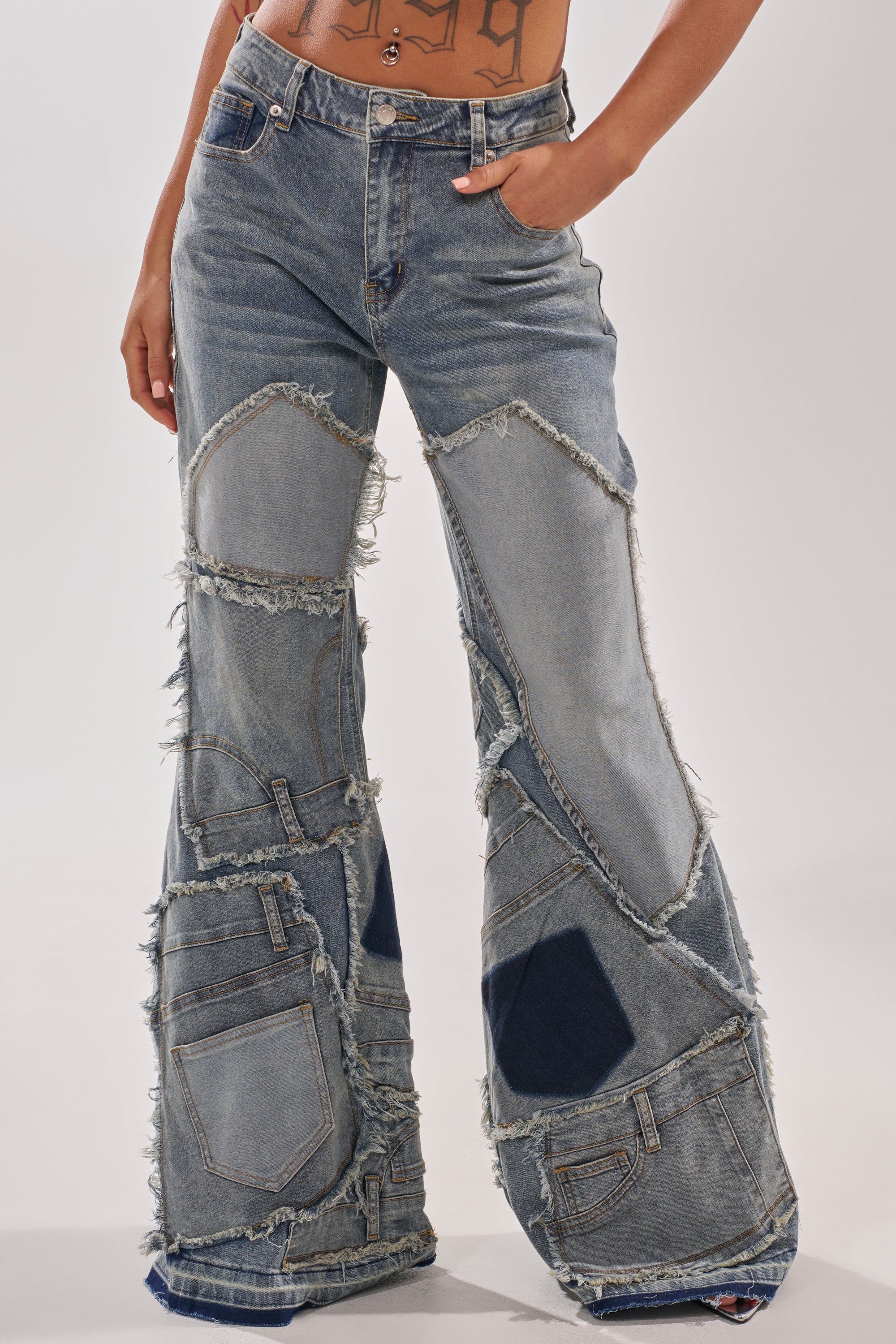 Wearing the ALL OVER ME DENIM PATCHWORK JEANS with high rise, frayed edges, and flared legs, a person stands with one hand in their pocket.