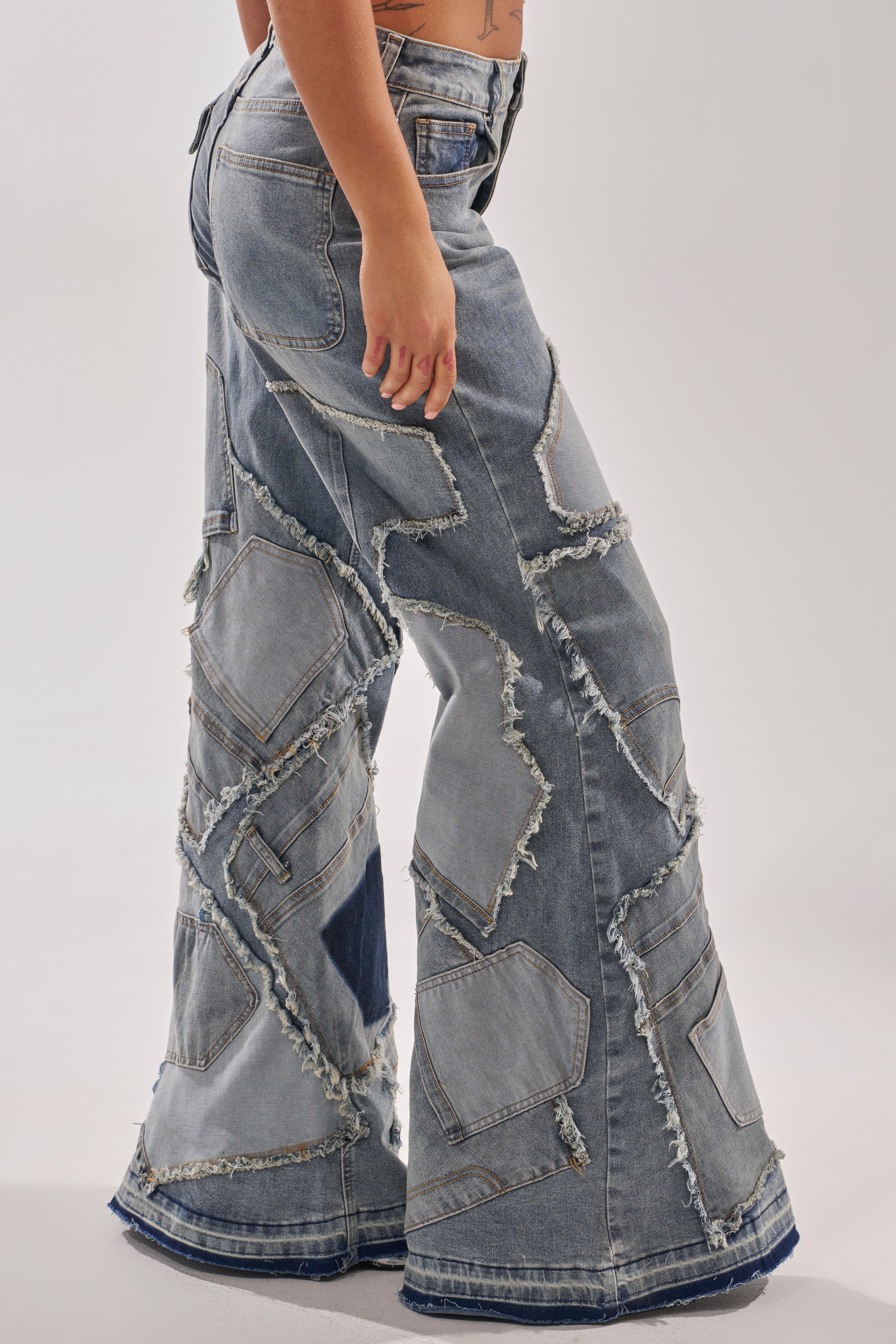 A person stands sideways against a plain background wearing ALL OVER ME DENIM PATCHWORK JEANS, featuring high rise and wide, frayed legs.