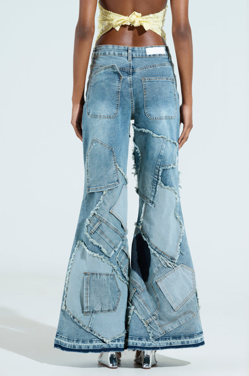 Wearing ALL OVER ME DENIM PATCHWORK JEANS with varying shades of flared denim, a person stands facing away. The high-rise fit pairs well with their yellow tied crop top, while visible stitching and frayed edges add detail. Shiny metallic shoes complete the look.