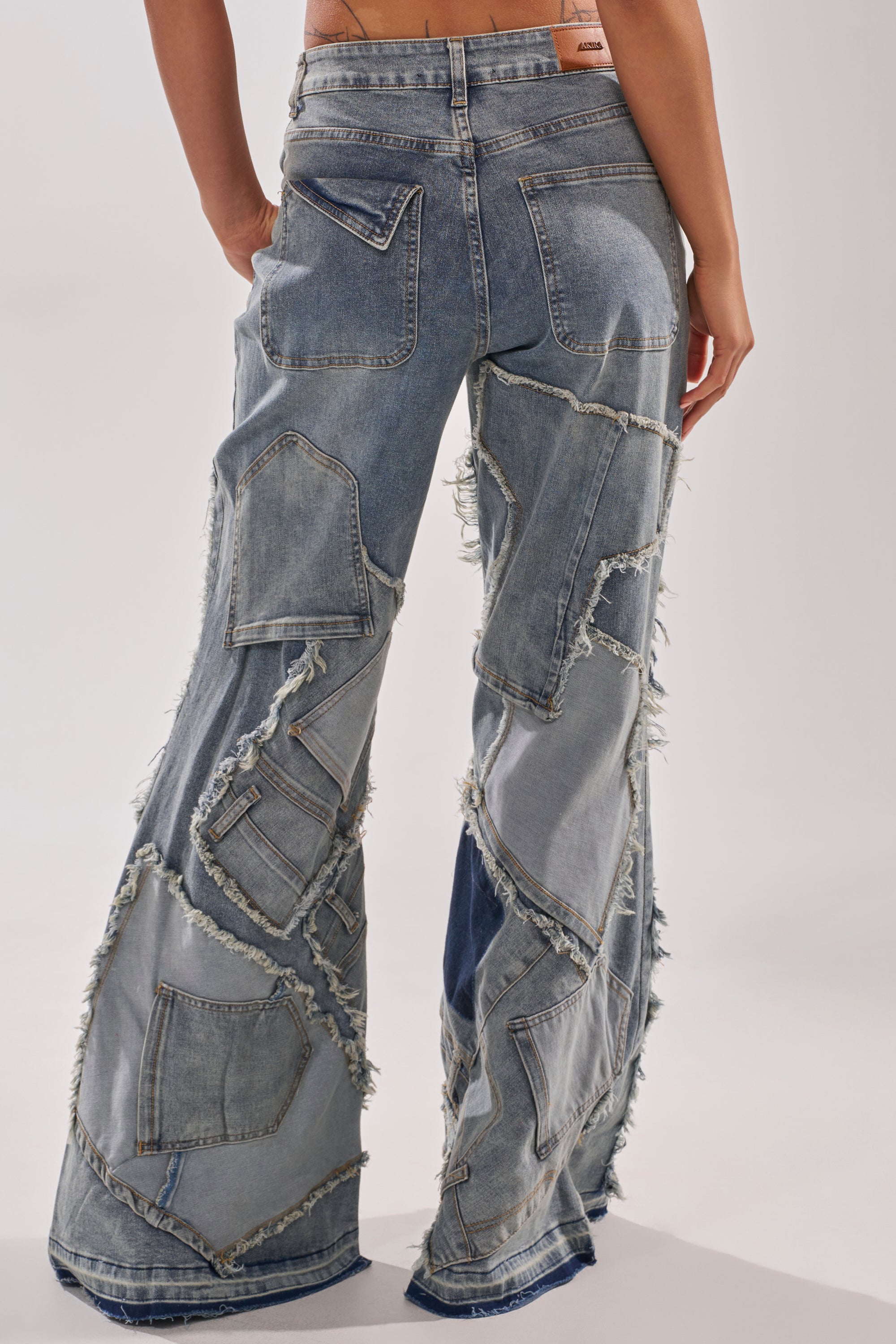 A person shown from the back wears ALL OVER ME DENIM PATCHWORK JEANS, featuring a high rise, flared leg, mixed denim shades, frayed edges, and exposed seams for a textured look.