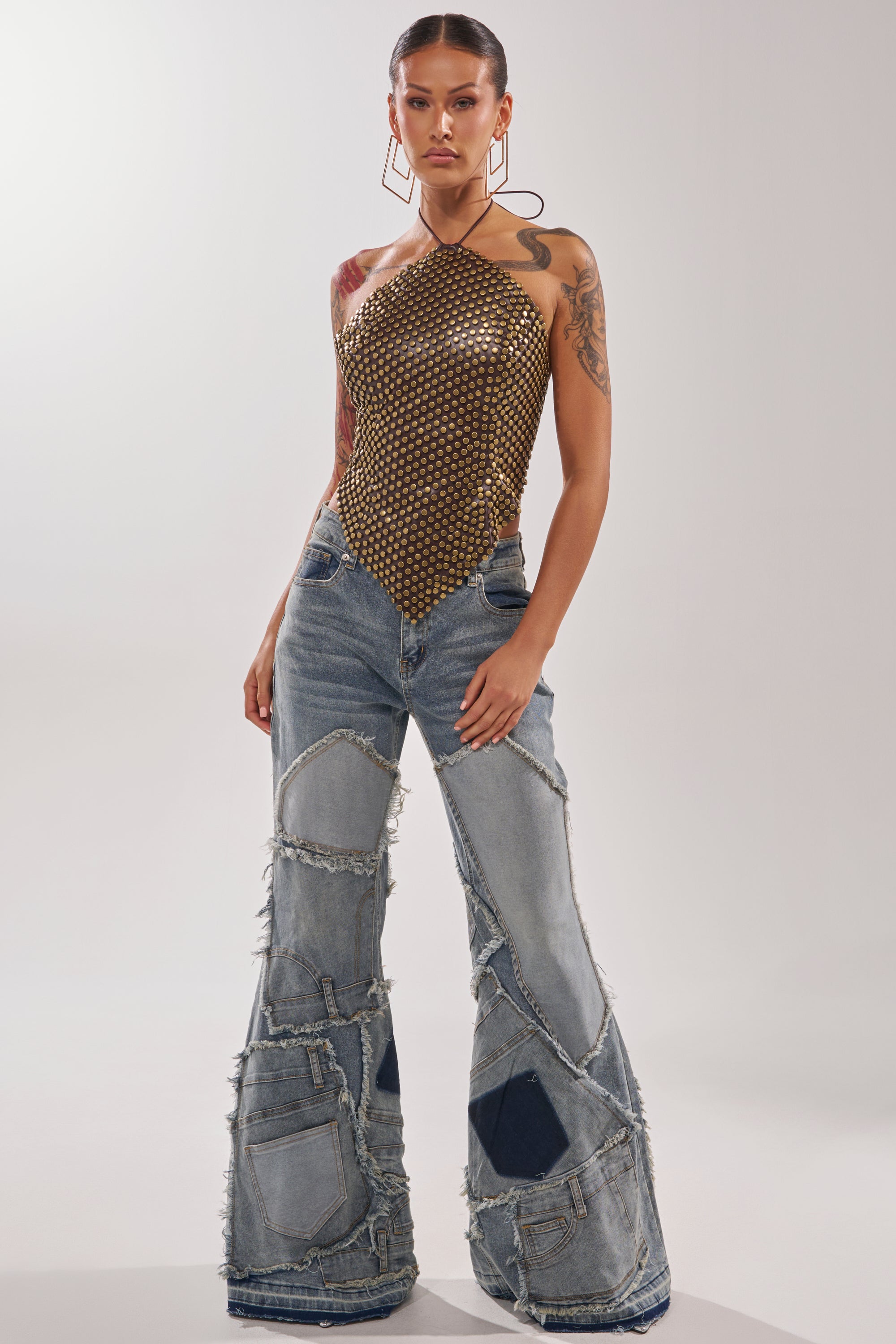 A woman poses confidently in ALL OVER ME DENIM PATCHWORK JEANS, styled with a gold halter-neck mesh top, bold geometric earrings, and visible tattoos, creating a striking look against a plain background.