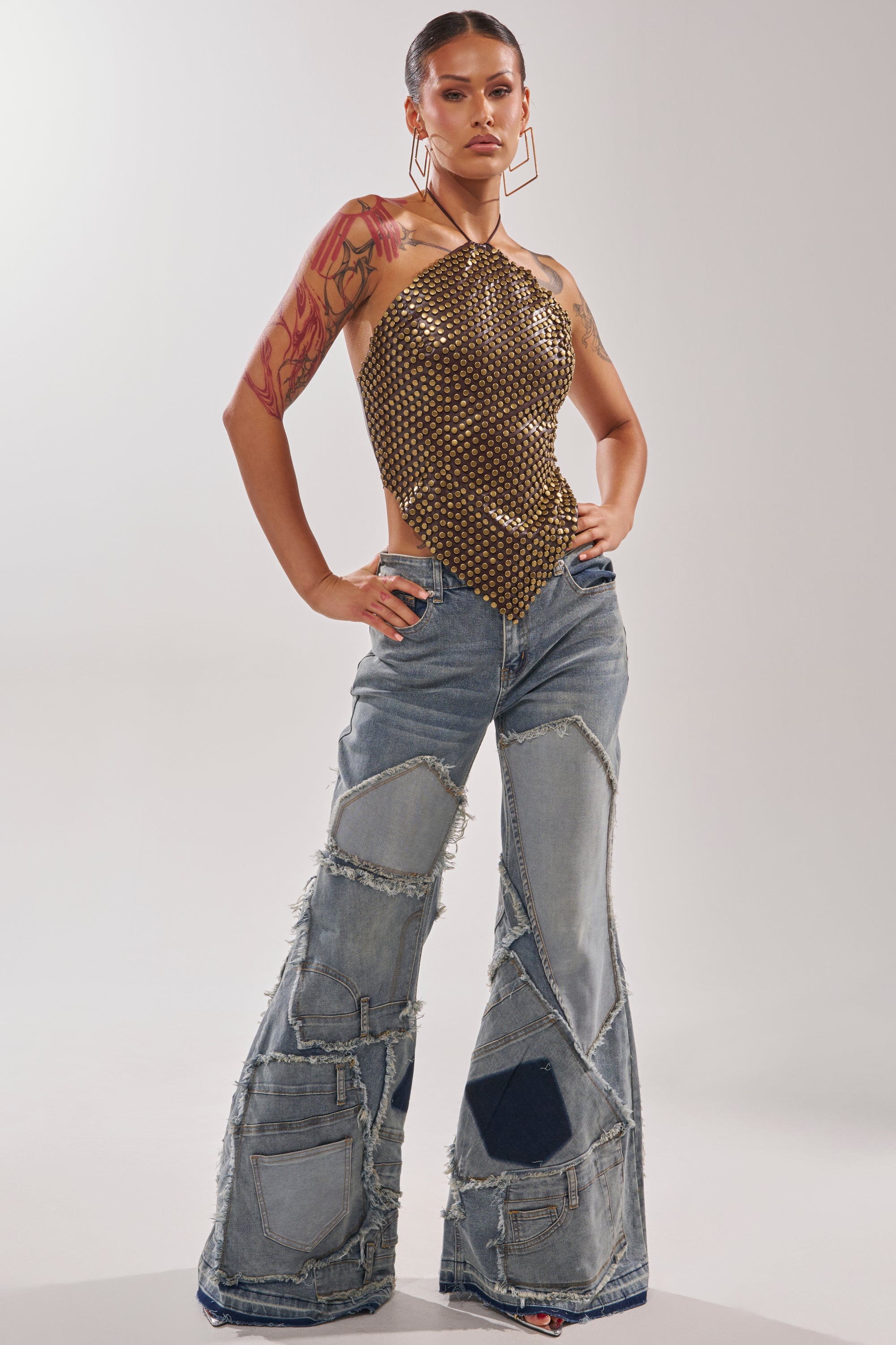 A person stands confidently in ALL OVER ME DENIM PATCHWORK JEANS with a halter-style metallic mesh top, featuring flared legs, raw hems, large pockets, geometric earrings, and visible arm tattoos.