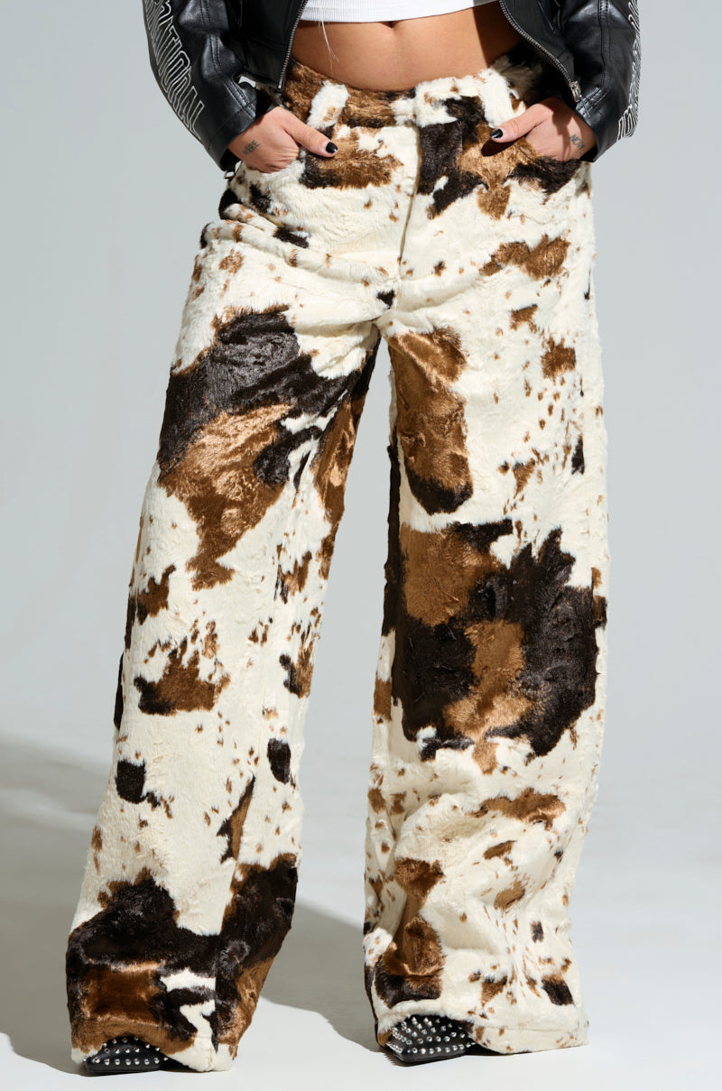 The individual is wearing the FEELING IT COW TROUSER, featuring wide legs and a high waist in a plush, textured cow print fabric. Paired with a black jacket, they have their hands comfortably tucked into the pockets of these stylish trousers.