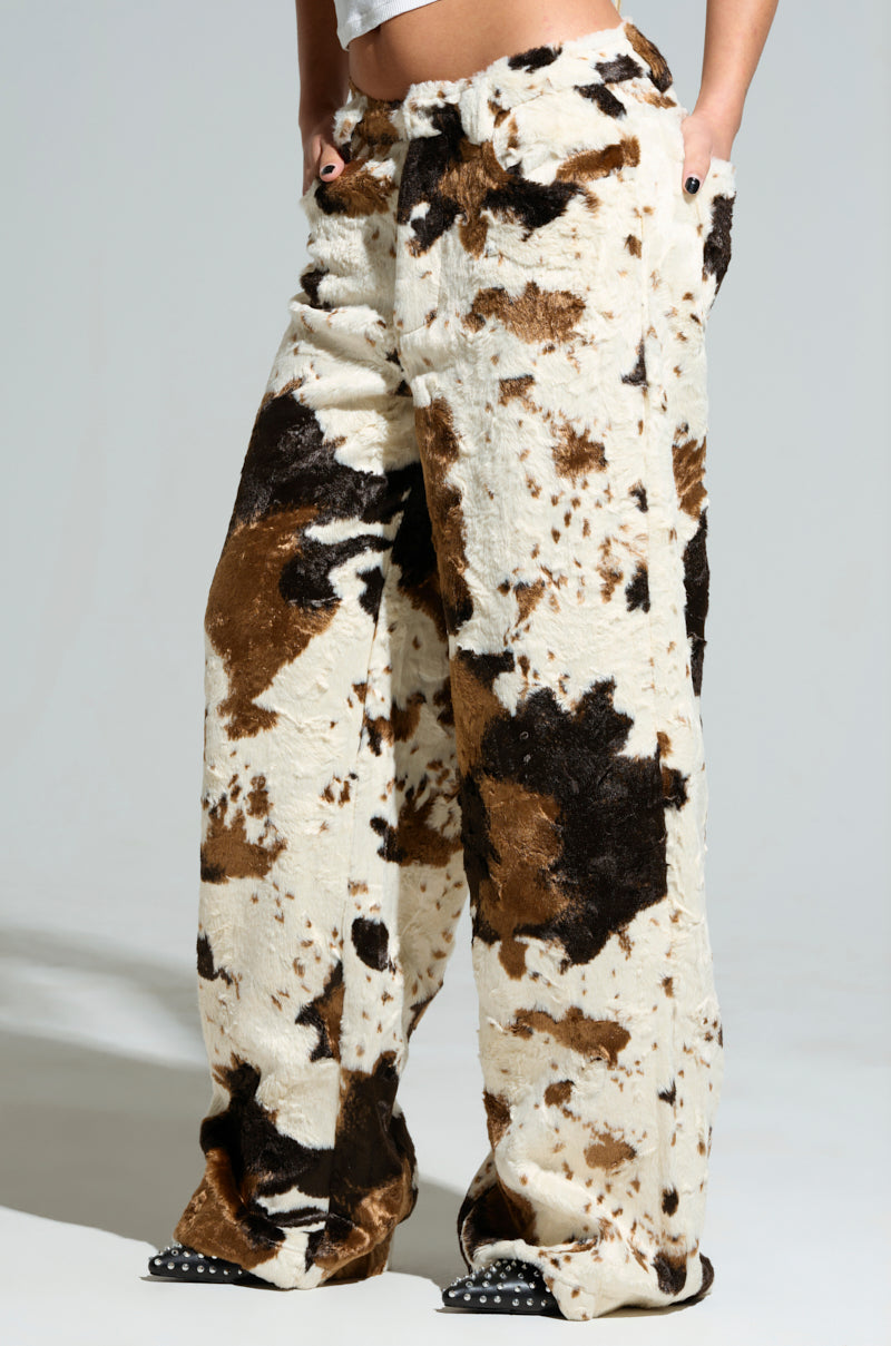 A person showcases the FEELING IT COW TROUSER, featuring wide-legged faux fur pants against a plain background. The predominantly white fabric is adorned with brown and black spots that closely mimic a cow pattern, as the individual casually places their hands in the pockets.