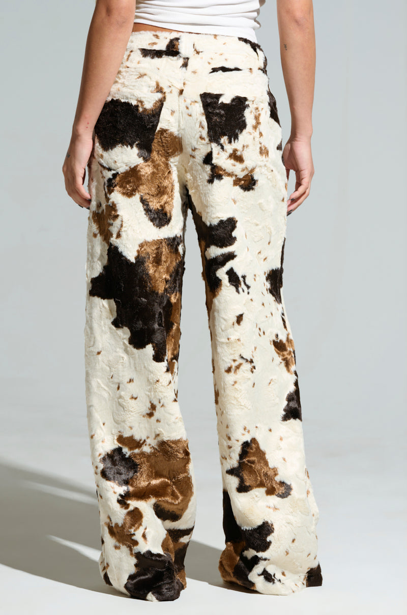 A person wearing the FEELING IT COW TROUSER, featuring a high-waisted design and cow print in black, brown, and white, stands with their back to the camera highlighting its full length and wide leg silhouette.